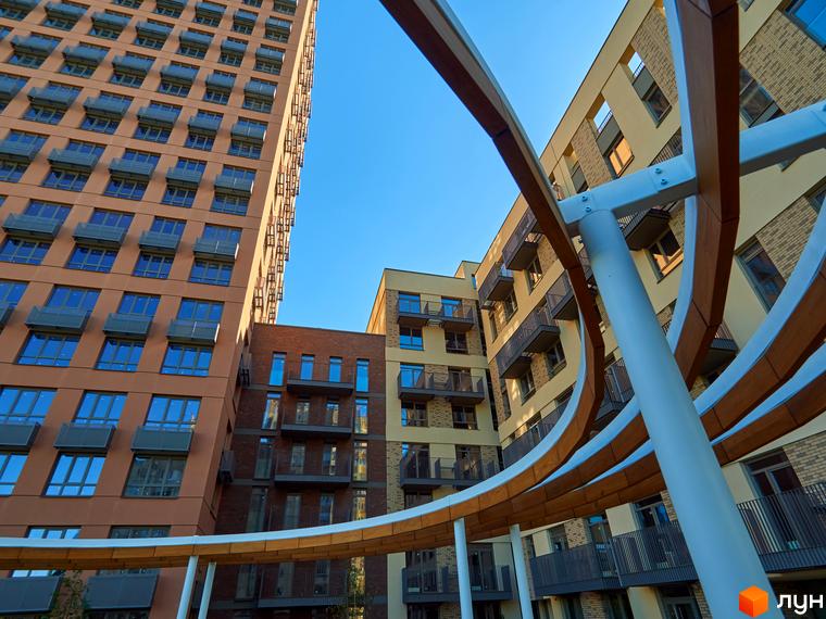 Будинок на Вавилових: високий коричневий будинок, світлі та цегляні будівлі з балконами. На передньому плані вигнута біла конструкція.