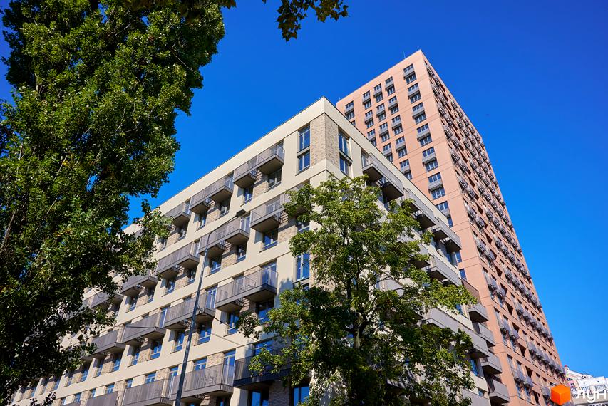 Висотна будівля житлового комплексу з балконами та панорамними вікнами на тлі блакитного неба. Поруч зелені дерева.