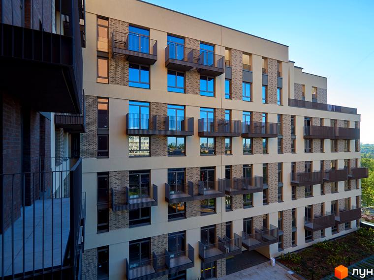 Багатоповерховий будинок з балконами та вікнами з відображенням неба. Біля будинку є невелика прибудинкова територія з рослинністю.