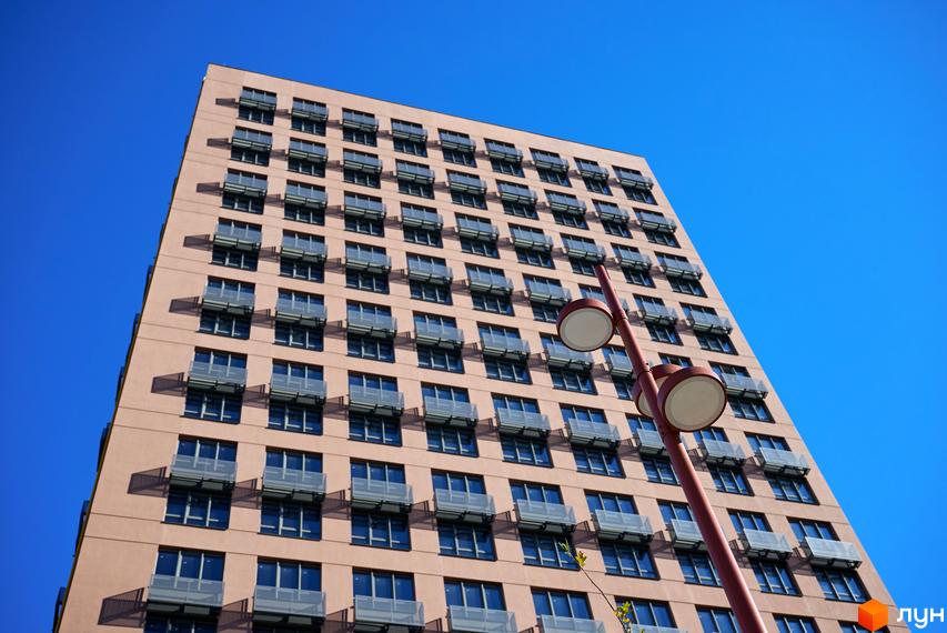 Фасад Будинку на Вавилових світло-коричневого кольору з численними вікнами та скляними огородженнями. На передньому плані видно вуличний ліхтар під яскравим синім небом.