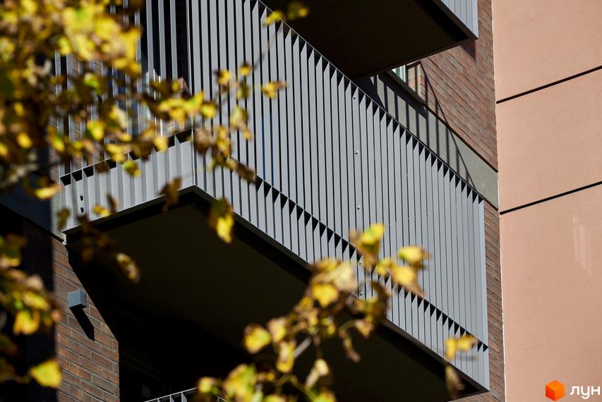 Фасад будинку ЖК Будинок на Вавилових з балконами та вертикальними ламелями, облицьований цеглою. Вид на прибудинкову територію з листям.