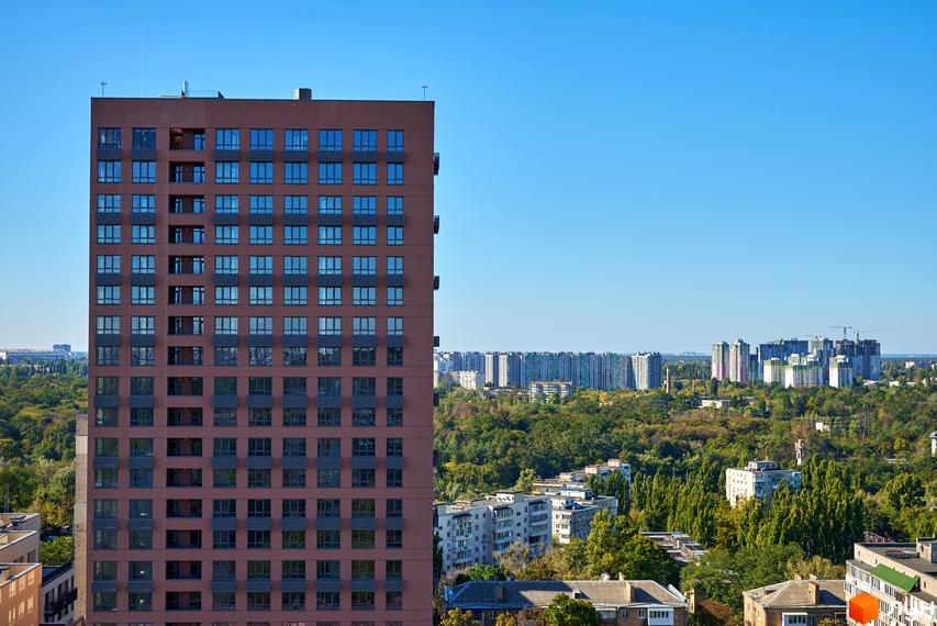 Висотна будівля житлового комплексу з великою кількістю вікон на фоні блакитного неба. Навколо будинку зелені дерева та інші будівлі.