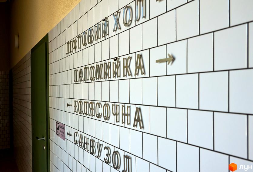 Напис з назвами приміщень на стіні з білої плитки. Зелені двері та коридор з білою плиткою.