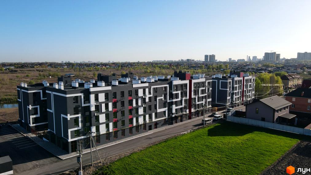 Зовнішній вигляд новобудови ЖК Рідний-2 з сірими фасадами та білими декоративними елементами. На прибудинковій території зелений газон та дорога.