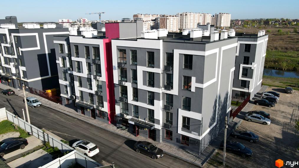 Житловий комплекс Рідний-2 з сірими фасадами та білими декоративними елементами, поруч розташовані припарковані автомобілі та зелена зона.