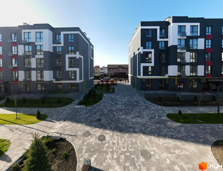 Зовнішній вигляд будівельного проєкту ЖК Рідний-2 з сірими та білими фасадами, що розташований на фоні блакитного неба. Прибудинкова територія з бруківкою та зеленими насадженнями.