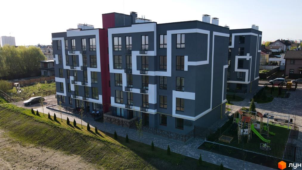 Зовнішній вигляд житлового комплексу Рідний-2 з сірими фасадами, білими декоративними елементами та червоною вставкою. На прибудинковій території розташований дитячий майданчик.