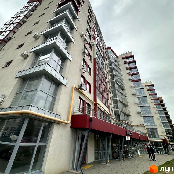 Житловий комплекс Європейський – багатоповерховий будинок з червоними та сірими елементами, скляними балконами та кондиціонерами. Поруч розташована бруківка та вхід до комерційних приміщень.