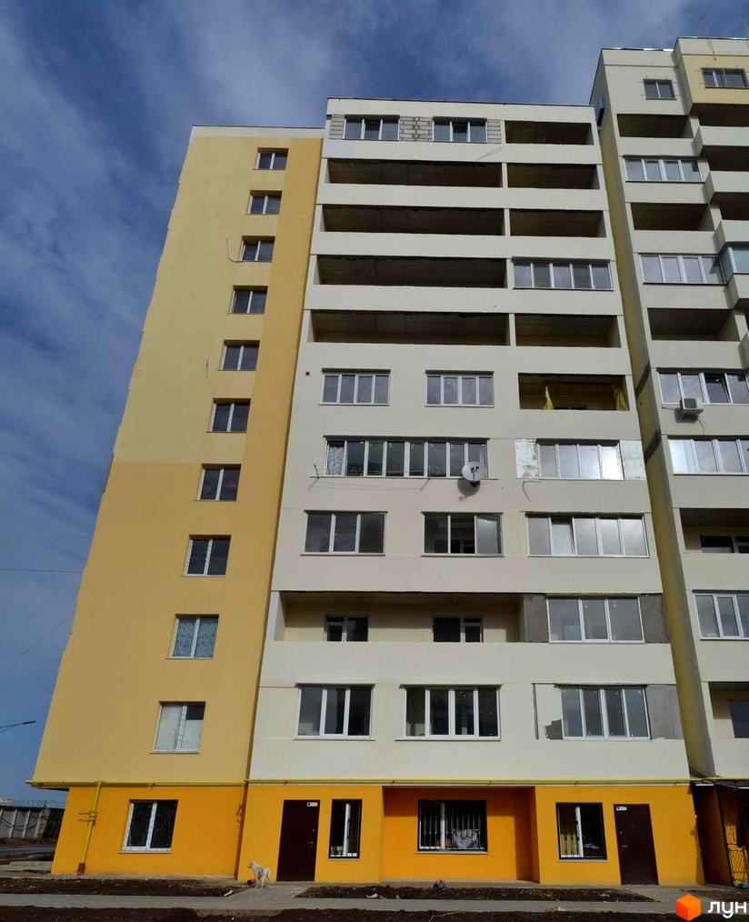Висотна будівля житлового комплексу з великою кількістю вікон та балконів. Фасад будинку виконаний у світлих тонах з жовтими акцентами.