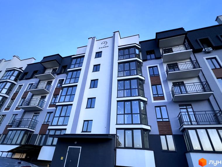 Зовнішній вигляд новобудови ЖК 9 Район з великою кількістю вікон та балконів. Фасад будинку світло-сірого кольору з темними елементами.