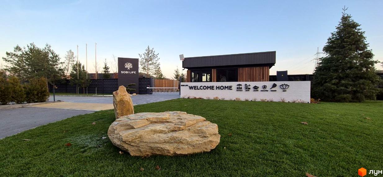 Вхід до Котеджного містечка Sobi Life з охоронним пунктом, великим білим знаком та зеленим газоном з каменями.