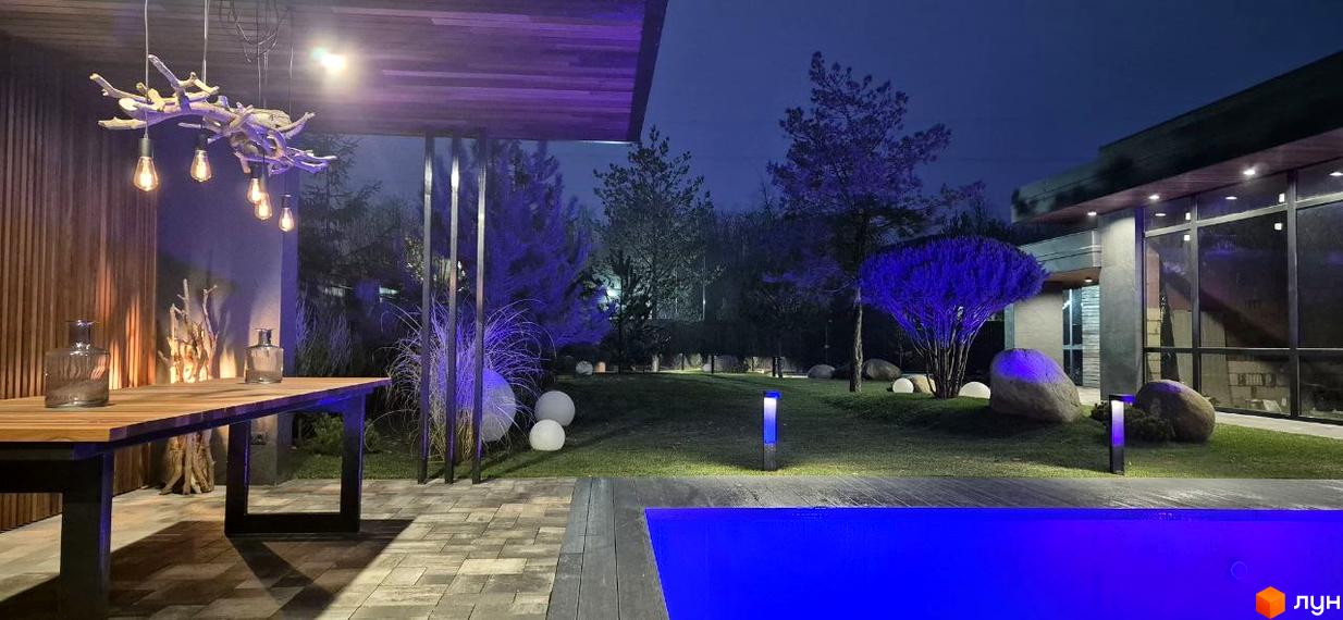 Котеджне містечко КМ Sobi Life з підсвіченим басейном та зоною відпочинку вночі. Навколо будинку зелений газон та дерева з фіолетовою підсвіткою.