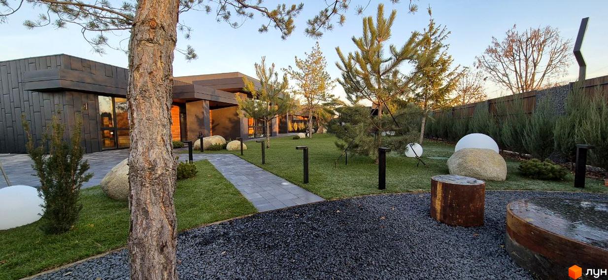 Будинок Котеджного містечка Sobi Life з темним фасадом та великими вікнами. На території зелений газон, доріжки, дерева та декоративні елементи.