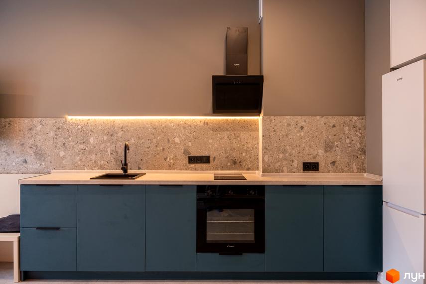 Сучасна кухня з синіми шафами, чорною раковиною та витяжкою. На стіні плитка з кам'яною текстурою та підсвічуванням.
