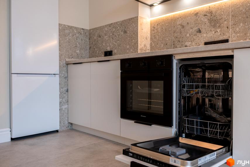 Сучасна кухня з вбудованою технікою: білий холодильник, чорна духова шафа та посудомийна машина. Сіра плитка на стінах та світла підлога.