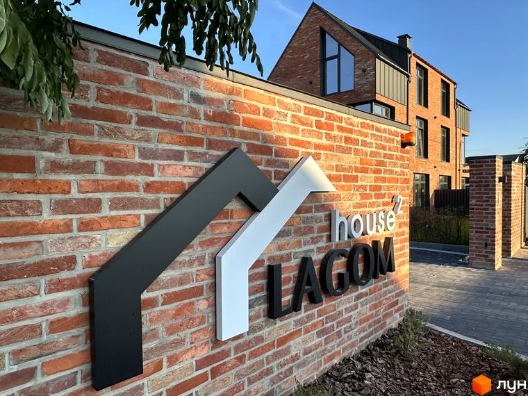 Зовнішній вигляд таунхаусів Lagom House з цегляним фасадом та логотипом комплексу. На передньому плані цегляна стіна з написом, позаду видно будинок та бруківку.