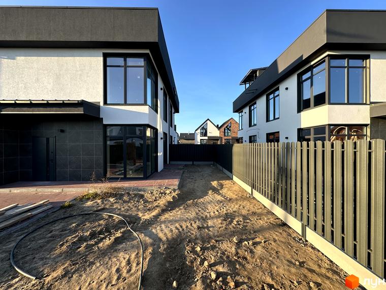 Таунхауси Lagom House з білою та чорною обробкою фасаду, великими вікнами та сірим парканом на тлі блакитного неба. Прибудинкова територія з піщаним покриттям та елементами ландшафтного дизайну.