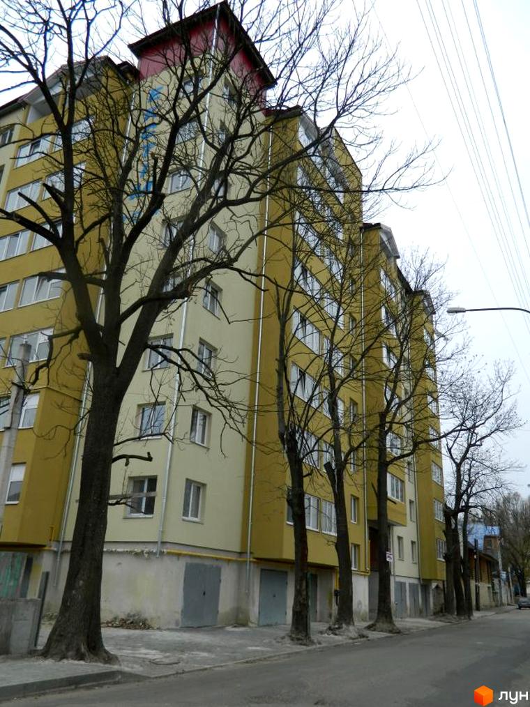 Зовнішній вигляд багатоквартирного будинку по вул. Ольги Кисілевської, 10. Житловий будинок з великою кількістю вікон та деревами вздовж тротуару.