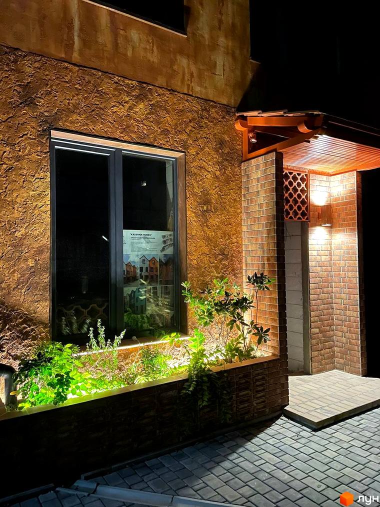 Зовнішній вигляд будинку КМ KADENIUK homes з цегляною кладкою та декоративною штукатуркою, освітленням та озелененням біля входу. Територія перед будинком вимощена плиткою.