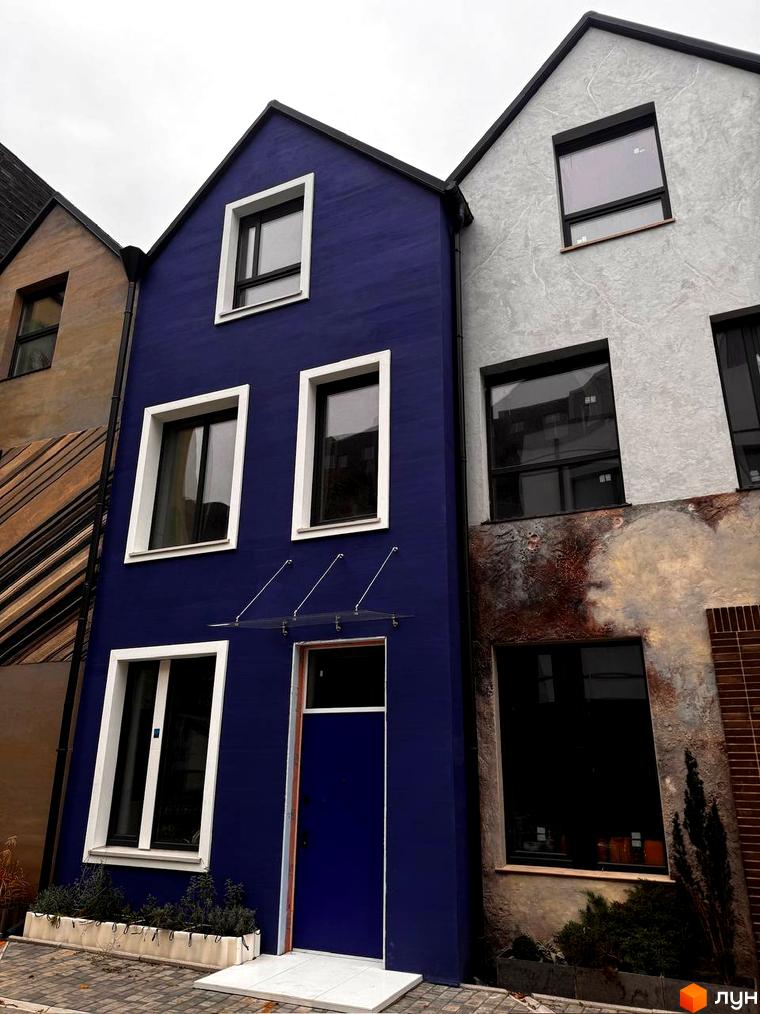 Фасад будинку КМ КАDЕНЮК homes з синім та сірим оздобленням, вікнами та дверима. Поруч розташовані інші будівлі та зелені насадження.