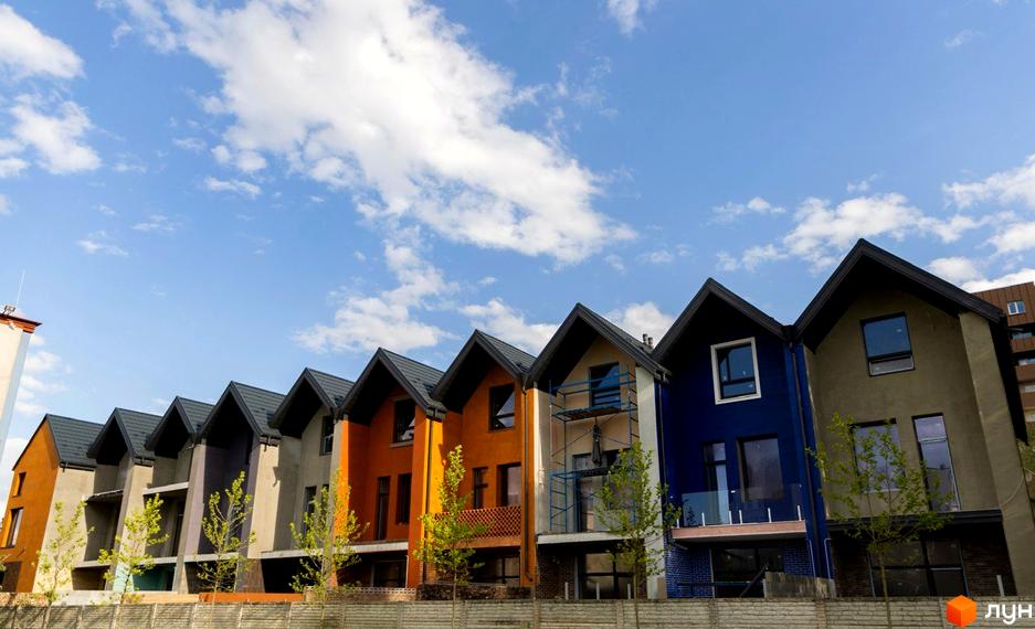 Котеджне містечко КМ КАDЕНЮК homes: ряд різнокольорових двоповерхових будинків з темними дахами під блакитним небом з хмарами. Перед будинками - зелені дерева та паркан.