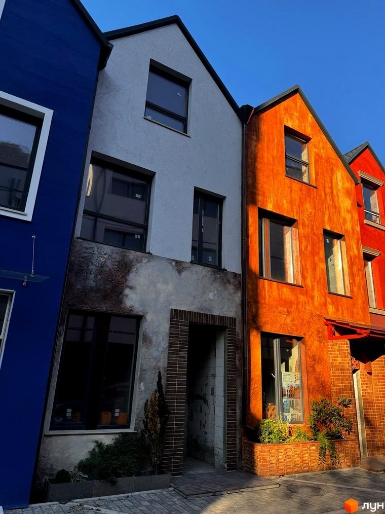 Фасади будинків КМ KADENYUK homes у яскравих кольорах: синій, білий, помаранчевий та червоний. Навколо будинків облаштовані клумби з рослинами та вимощені доріжки.