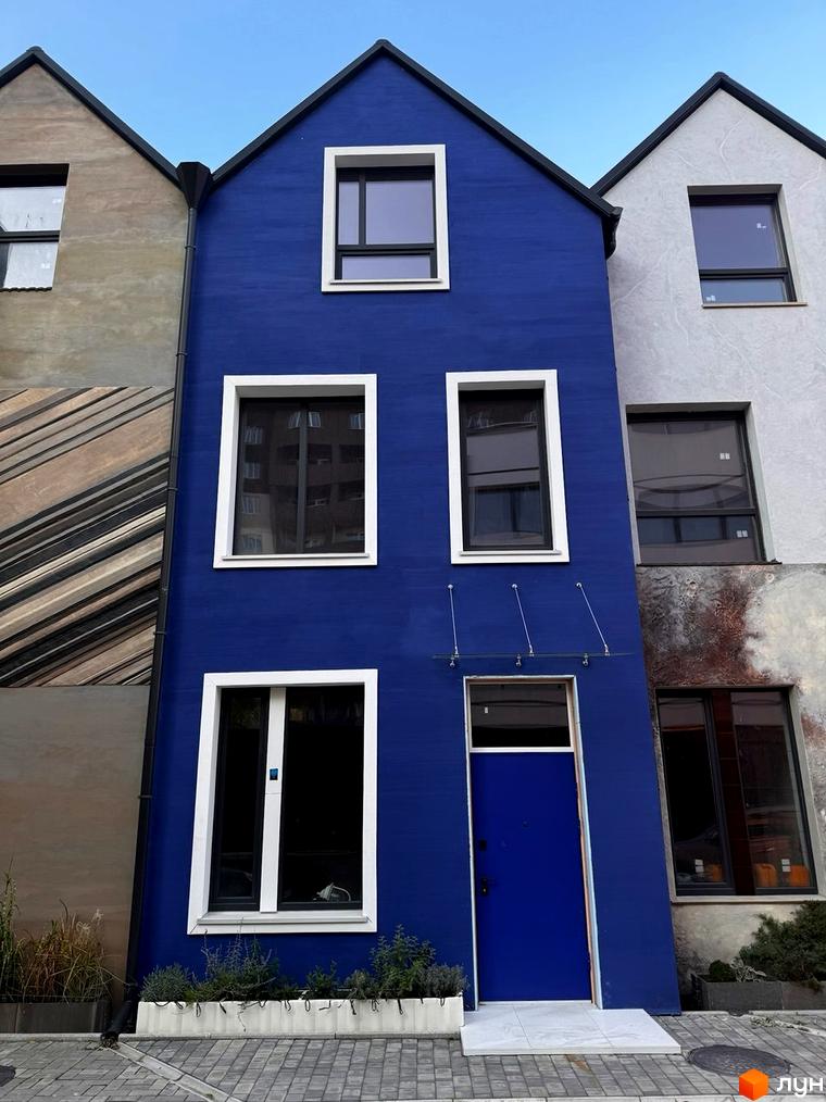 Фасад будинку житлового комплексу КМ KADENIUK homes з яскраво-синьою стіною та вікнами. Перед будинком розташована прибудинкова територія з рослинами.