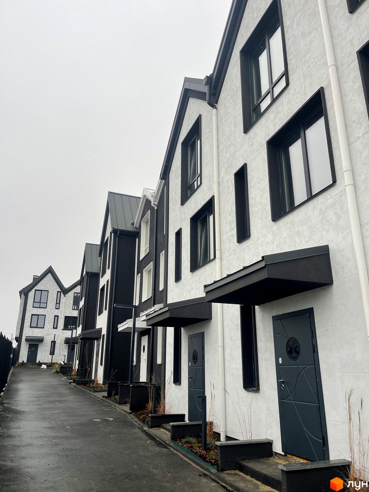 Таунхаус New Smart 15 - сучасний житловий комплекс з біло-чорними фасадами та стильними дверима, розташований на схилі з асфальтованою дорогою.