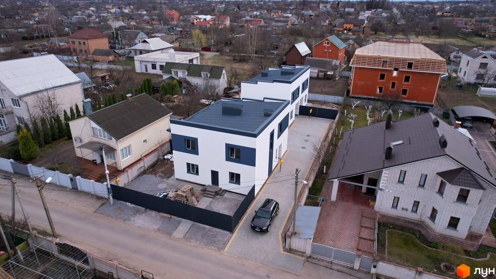 Зображення житлового будинку з білими стінами та синіми акцентами, розташованого на території з бруківкою, поруч з іншими будинками в околицях.