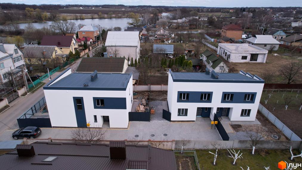 Дуплекси Флагман Хутори: двоповерхові будинки білого кольору з темно-синіми вставками та сірими дахами, розташовані на доглянутій прибудинковій території з бруківкою та парканом.