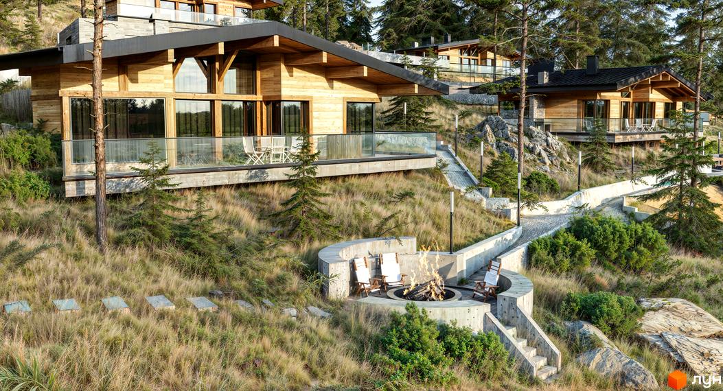 Котеджне містечко Veres Hill з дерев'яними фасадами та панорамними вікнами, розташоване на схилі серед дерев. На прибудинковій території облаштований бетонний майданчик з вогнищем та зручними кріслами.