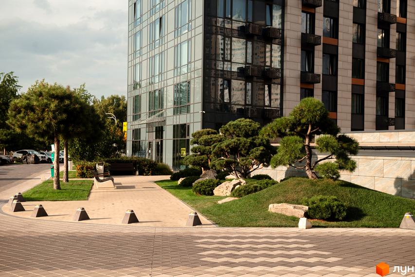 Житловий комплекс Метрополіс з фасадом зі скла та каменю. Навколо комплексу доглянута територія з деревами та зеленими насадженнями.