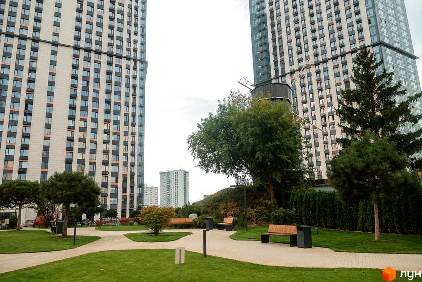 Житловий комплекс Метрополіс: високі будинки зі світлим фасадом та численними вікнами. Прибудинкова територія має зелені газони та зони відпочинку з лавками.