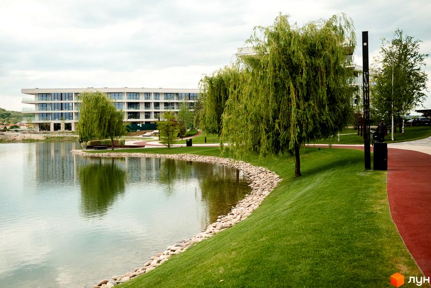 Сучасний житловий комплекс Park Lake City з великою зеленою територією та озером. Будівля має багато вікон та балконів, що виходять на воду та парк.