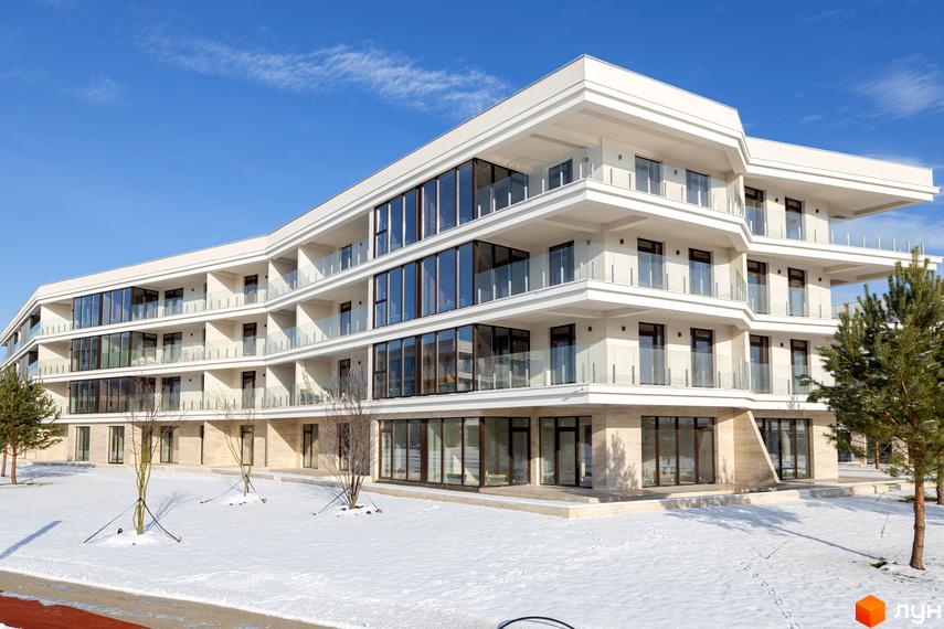 Житловий комплекс Park Lake City із скляними балконами та великими вікнами на тлі блакитного неба. Навколо будівлі засніжена територія з молодими деревами.