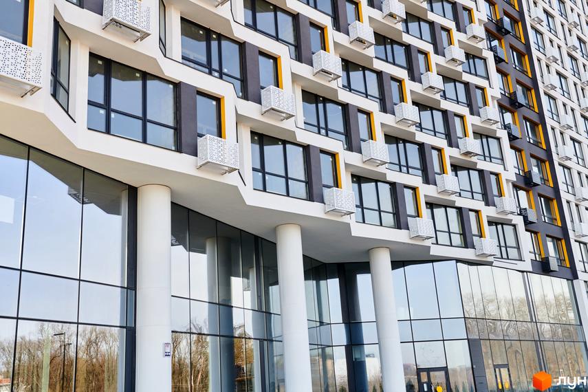 Новобудова LUCKY LAND з багатоповерховим фасадом у білих, сірих та жовтих тонах, панорамними вікнами та скляним першим поверхом. Поруч видно дерева.