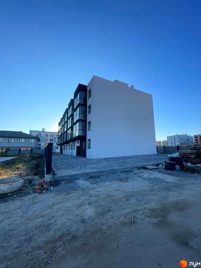 Білий багатоквартирний будинок з темними вікнами та балконами. Перед будинком ґрунтова ділянка та бруківка.