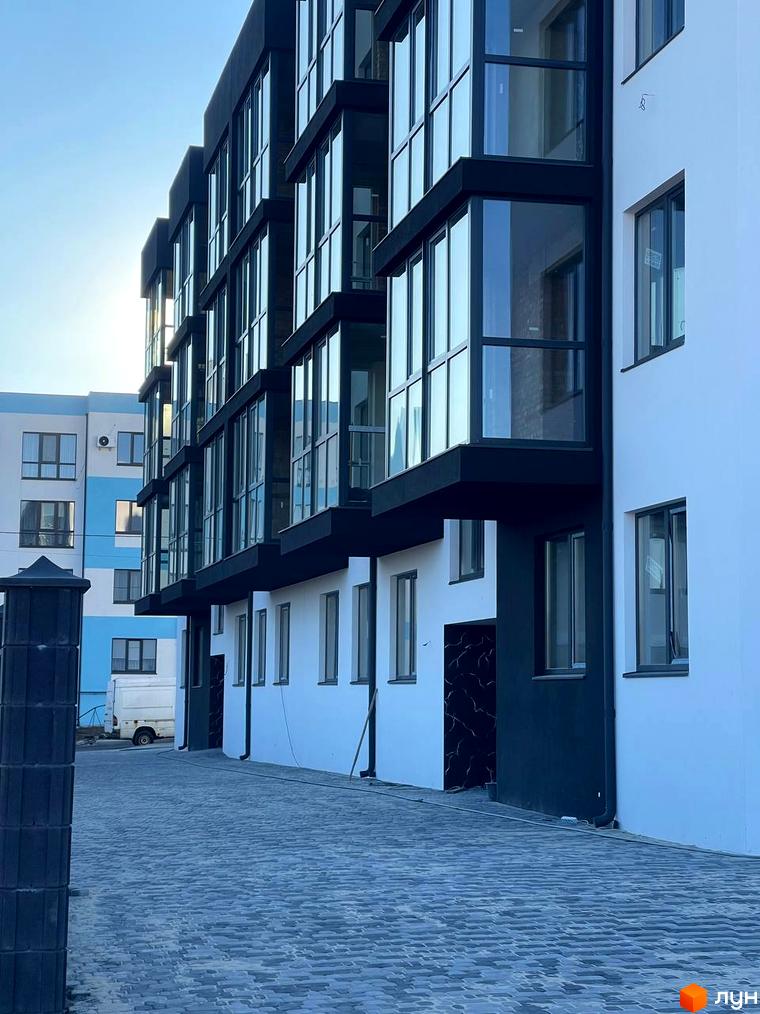 Білий багатоквартирний будинок з темними віконними рамами та заскленими балконами. Перед будинком викладена сіра бруківка.