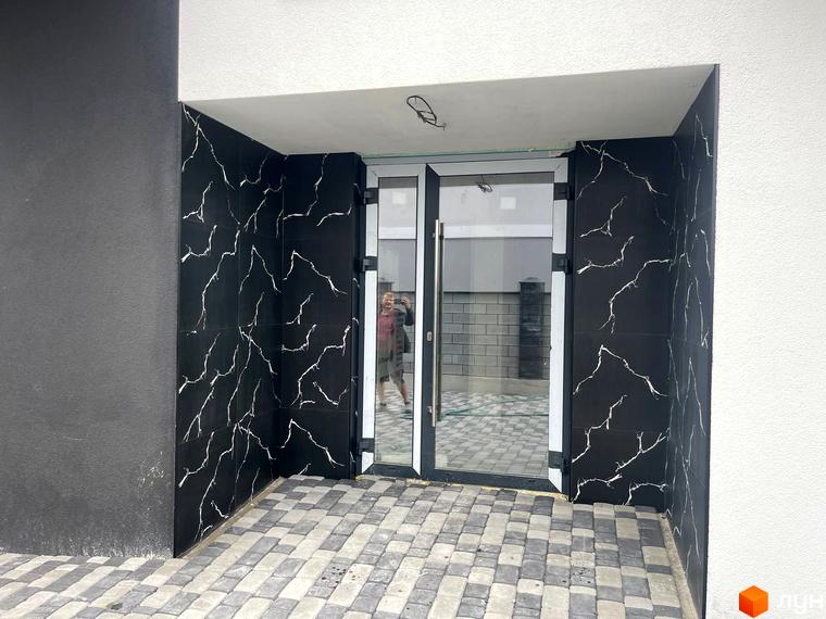 Вхід до новобудови на Фабричній з чорними стінами, оздобленими білим візерунком, та скляними дверима. Прибудинкова територія вимощена сірою бруківкою.