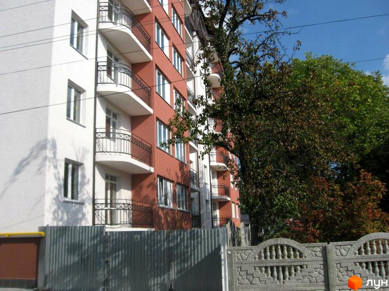 Зовнішній вигляд багатоповерхового будинку з балконами. Житловий комплекс розташований біля зелених насаджень та має огорожу.