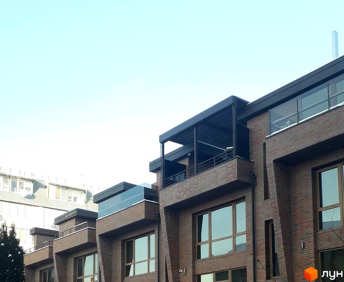 Фасад будинку житлового комплексу на вул. Вільхова, 7в, з цегляним оздобленням та скляними балконами. Навколо видно дерева та сусідні будівлі.