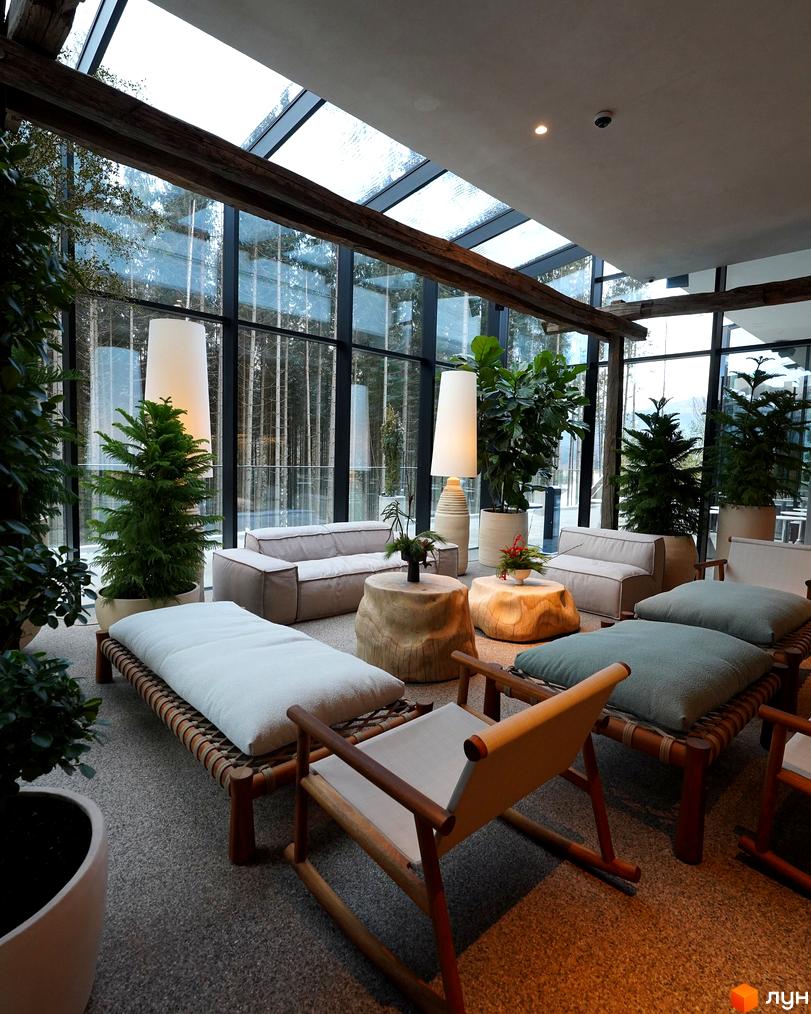 Інтер'єр лобі житлового комплексу Mountain Residence з великими панорамними вікнами, що виходять на ліс. Затишна зона відпочинку з диванами та кріслами.