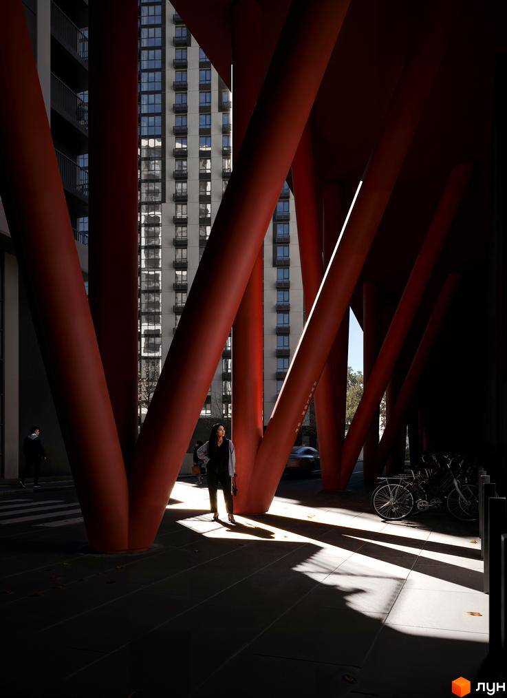 Житловий комплекс Avalon Up з червоними опорами та фасадом з вікнами. Поруч розташовані велосипеди та люди.