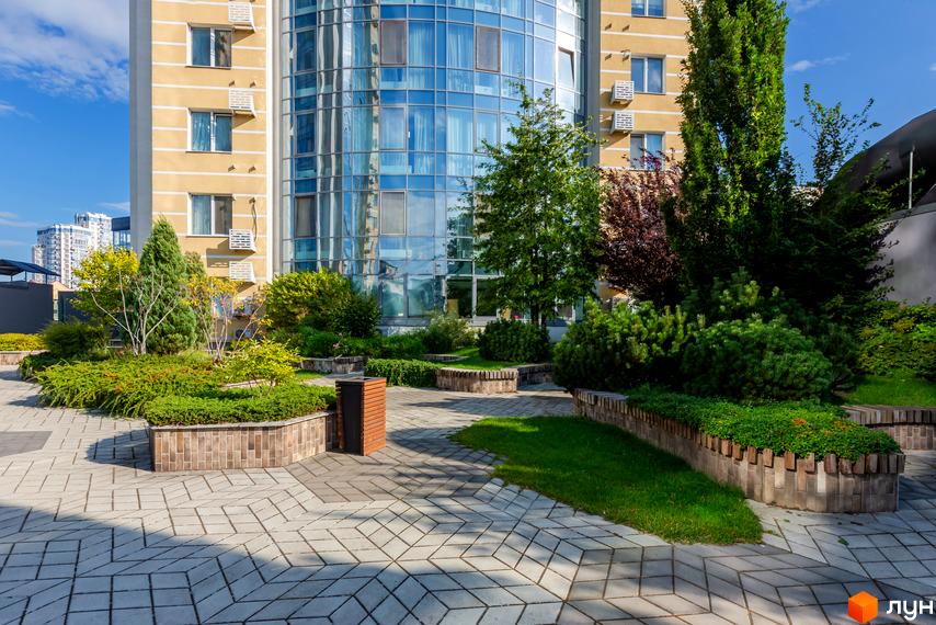 Будівля Житлового комплексу Smart Plaza Polytech з жовтим фасадом та вигнутою скляною частиною. Прибудинкова територія озеленена деревами, кущами та має бруковані доріжки.