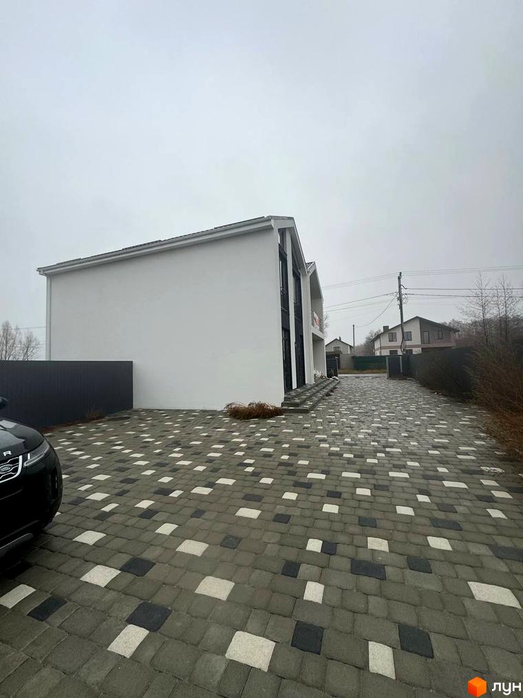 Сучасний білий дуплекс Family House з панорамними вікнами та бруківкою на подвір'ї. Територія біля будинку засаджена деревами та кущами.