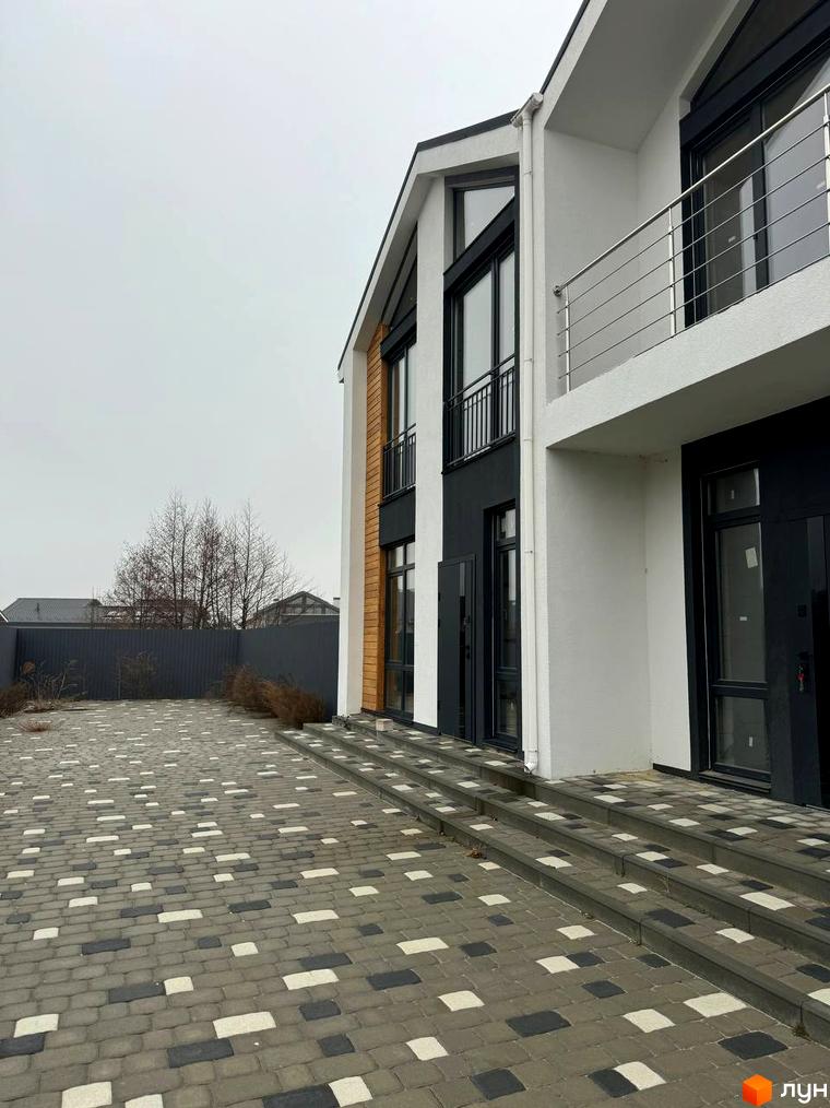 Фасад дуплексу Family House з білими та темними елементами, дерев'яним оздобленням та балконом. Перед будинком вимощена територія з різнокольорової бруківки.