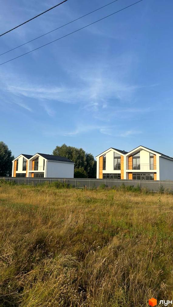 Два сучасних дуплекси Family House з білими фасадами та дерев'яними елементами на тлі блакитного неба та зелених дерев. Навколо будинків невисокий паркан та поле з високою травою.