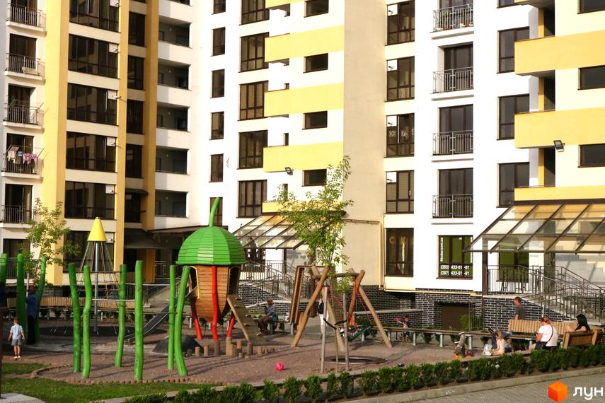 Житловий комплекс Lystopad: білі та жовті фасади, балкони з металевими огорожами. Дитячий майданчик з гіркою, гойдалкою та пісочницею.