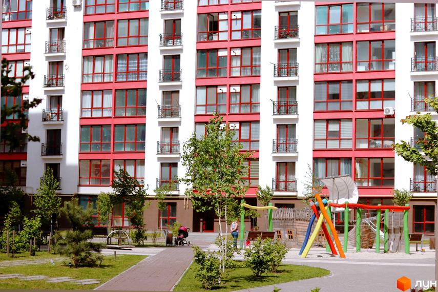 Зображення фасаду багатоповерхового будинку ЖК Містечко Козацьке з червоними балконами та вікнами. На передньому плані дитячий майданчик та зелені насадження.