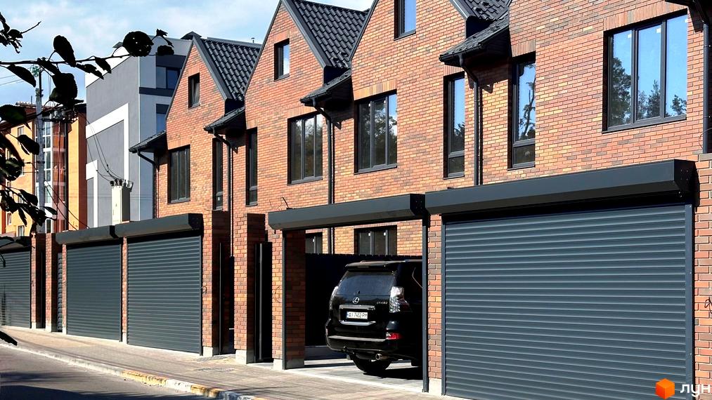 Цегляний будинок з гаражними воротами та чорним авто. На задньому плані видно інші будівлі та блакитне небо.